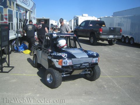 1990 Ford Bronco - black-and-silver No. 98 off-road race truck with a helmeted driver sits among RVs and trailers in a paddock.