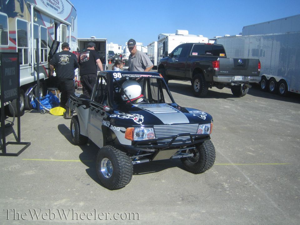 Black and silver number 98 off-road race truck parked among trailers in a paddock