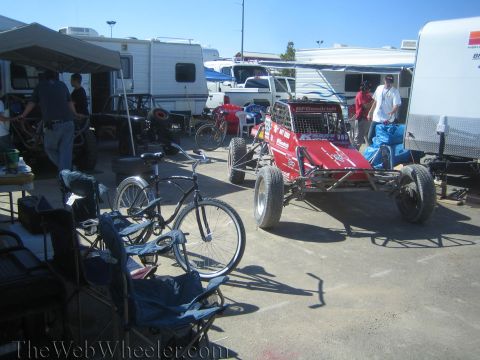1990 Ford Bronco - red BFGoodrich-liveried tube-chassis off-road racer sits in a crowded RV paddock alongside parked bicycles and lawn chairs.