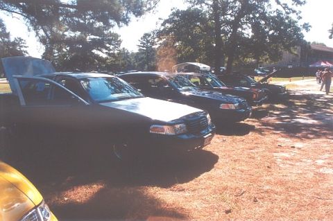 2002 Ford Crown Victoria - A line of sedans, including a dark 2002 Ford Crown Victoria with its hood and driver door ajar, are parked outdoors.
