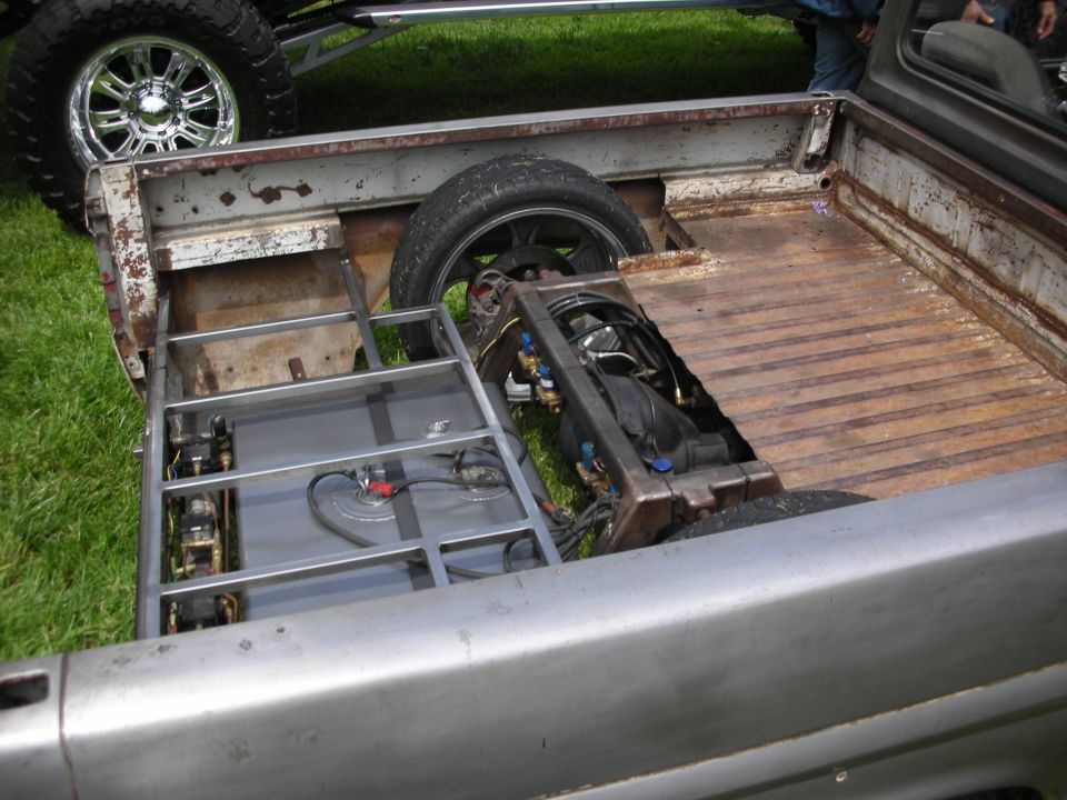 Custom pickup truck bed showing fuel tank, suspension, and spare tire. Another vehicle's wheel is visible