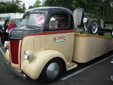 1992 Ford Crown Victoria - A vintage tow truck painted beige and black carries a hot rod on its trailer at a car show.