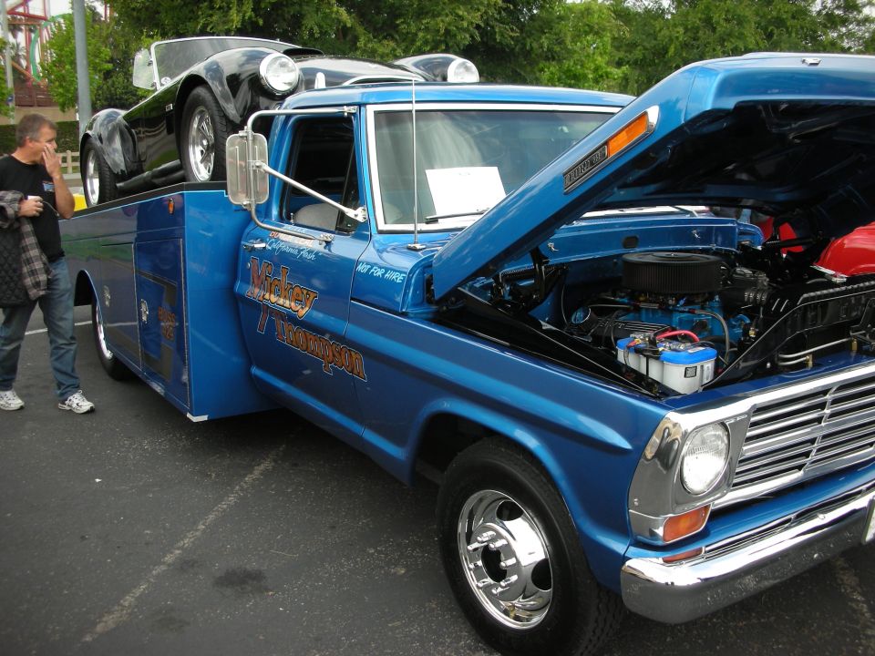 Blue tow truck with hood open, carrying a black sports car on its bed. Man stands nearby