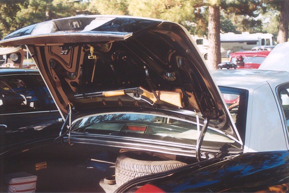 Car trunk open, showing spare tire and some storage. Another car is visible to the left. RVs and a red car are in the background