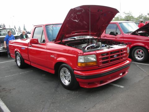 1992 Ford Crown Victoria - A red 1993 Ford F-150 Lightning pickup truck is displayed with its hood open, revealing the engine bay. Another similar truck is parked alo…