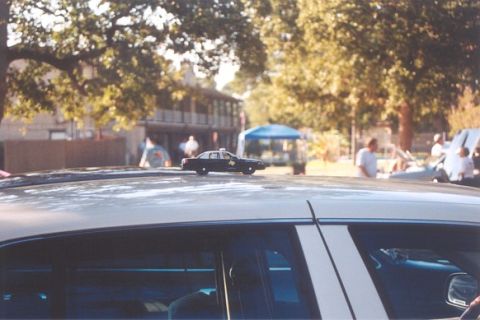 2002 Ford Crown Victoria - A toy police car is placed on the roof of a vehicle, with a building and trees visible in the background.
