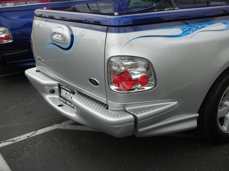 Rear of a silver Ford F-150 Lightning pickup with blue flame graphics. Another blue truck is partially visible