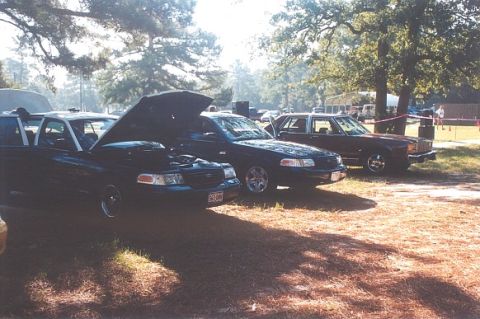 2002 Ford Crown Victoria - A dark 2002 Ford Crown Victoria is parked with its hood open, alongside other vehicles in an outdoor setting.