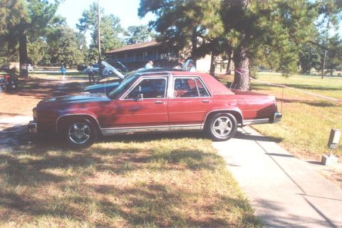 2002 Ford Crown Victoria - Red sedan parked on a sidewalk with trees and a building in the background.