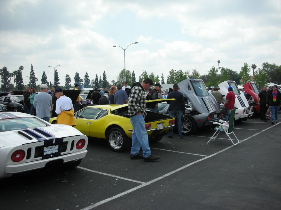 Cars parked in lot, people milling around. White car in foreground, yellow sports car, then several gray, white, and red cars with hoods up