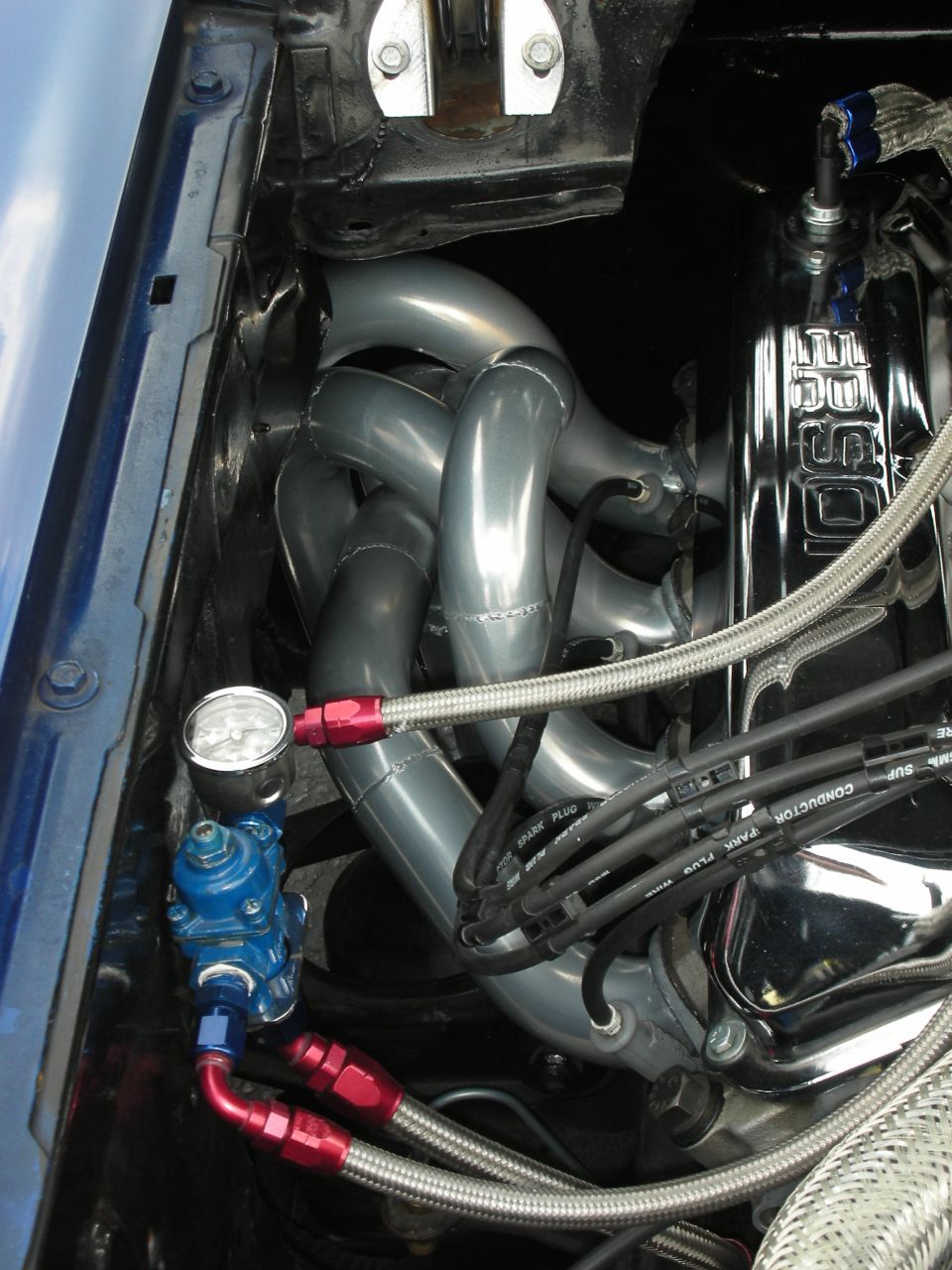 Close-up of a car's engine bay showing shiny exhaust headers, a blue fuel pressure regulator with red fittings, and spark plug wires