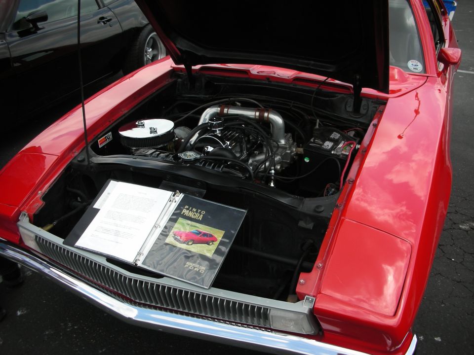 Red car engine bay with open hood, showing engine and a binder with 'PINTO PANGRA' on it. Another car is visible in the background
