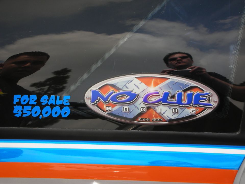 Car window with 'FOR SALE $50,000' sticker and 'NO CLUE RACING SINCE 2000' oval sticker. Reflections of people and sky are visible