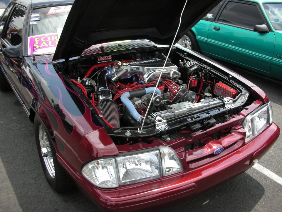 Modified 1991 Ford Mustang engine bay with custom red hoses and chrome parts. Green Mustang in background