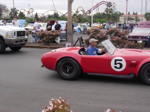1992 Ford Crown Victoria - A man drives a red convertible sports car with a large number 5 on its side, at an outdoor car show with a roller coaster in the background.