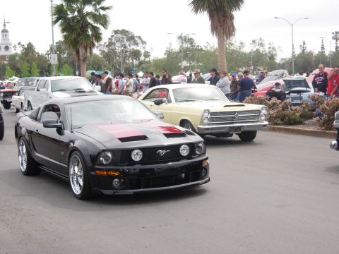 1992 Ford Crown Victoria - A black Ford Mustang with red hood stripes is seen at a car show, with other vehicles and spectators in the background.