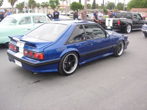 1992 Ford Crown Victoria - Blue Ford Mustang hatchback with custom wheels and racing stripes parked at an outdoor car show.