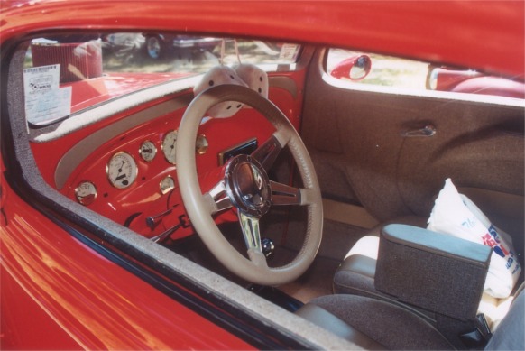 Interior of a red hot rod with a tan steering wheel, custom gauges, and cloth seats. A white bag is on the passenger seat