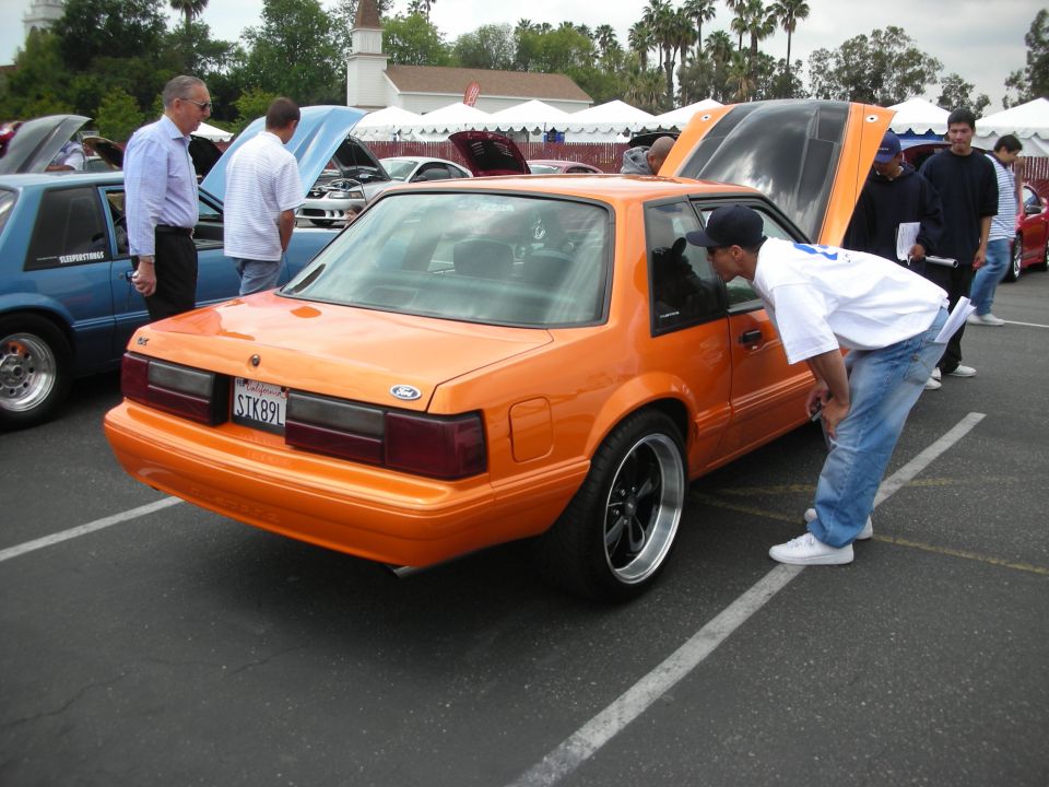 Orange Ford Mustang coupe, rear view, with a guy looking in the window. Another blue car is parked next to it