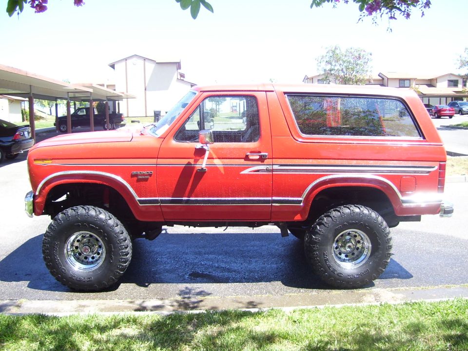 Side view of a red lifted 1986 Ford Bronco with chrome wheels and off-road tires parked outside