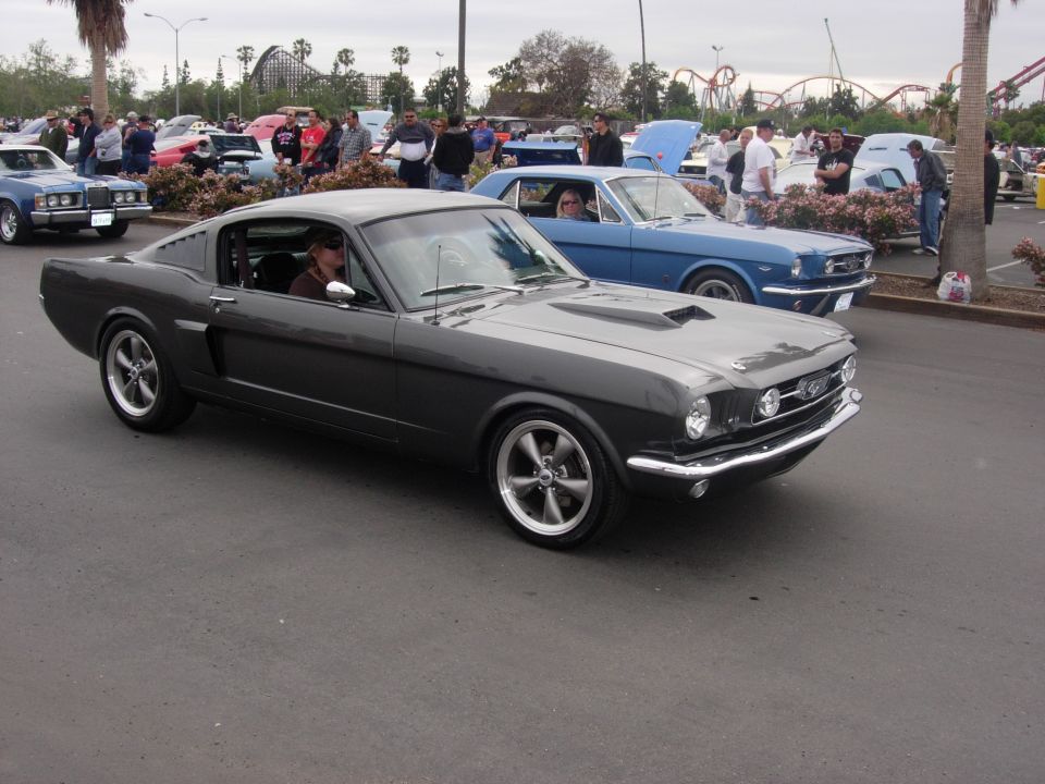 Dark gray 1965 Ford Mustang Fastback with aftermarket wheels, parked next to a blue classic car