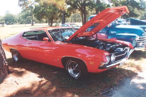 2002 Ford Crown Victoria - A red 1970s muscle car is parked with its hood open, revealing the engine, with a blue truck visible behind it.