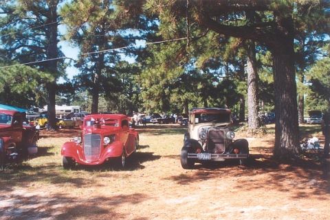 2002 Ford Crown Victoria - A gathering of classic vehicles parked outdoors under the shade of large trees.