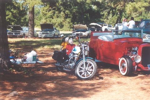2002 Ford Crown Victoria - A motorcycle with two figures is parked next to a red hot rod at an outdoor gathering with several other vehicles.