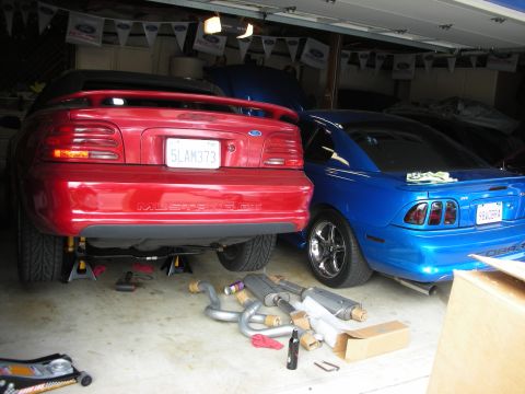 1998 Ford Crown Victoria - red Ford Mustang GT and a blue Mustang Cobra sit in a Ford-pennant-decorated garage with exhaust components laid out on the floor.