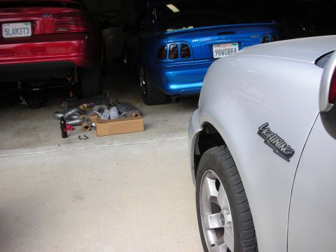 1998 Ford Crown Victoria - Red and blue Ford Mustangs sit in a garage next to a silver vehicle wearing a Lightning badge, with exhaust parts in a box on the floor.