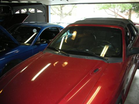 1998 Ford Crown Victoria - maroon convertible with a black soft top sits nose-out in a garage next to a blue car, light reflecting off its hood.