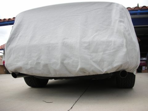 1998 Ford Crown Victoria - vehicle sits fully draped under a gray fitted car cover on a paved driveway, with exhaust tips and tires peeking out below.