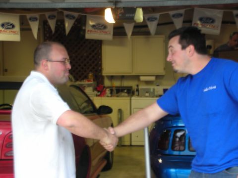 1998 Ford Crown Victoria - Two men shake hands in a garage decorated with Ford Racing pennants, with cars and laundry appliances in the background.