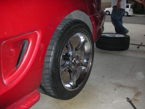 1998 Ford Crown Victoria - chrome five-spoke wheel and low-profile tire on a red car, with a spare wheel and tools lying on the driveway behind.