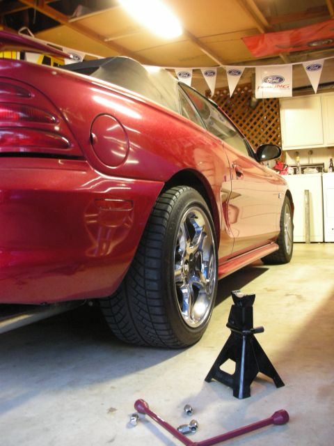 1998 Ford Crown Victoria - side of a red Ford Mustang convertible rests on a jack stand with a chrome five-spoke wheel, a lug wrench and nuts on the floor.