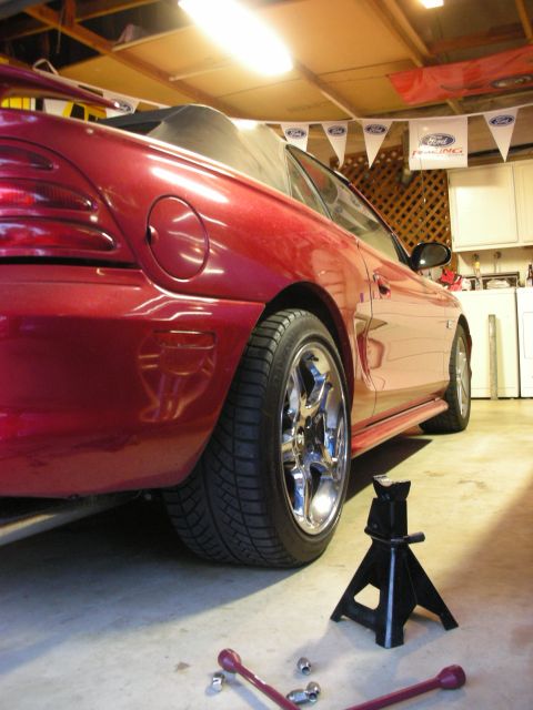 1998 Ford Crown Victoria - Low rear-quarter view of a red convertible coupe on a chrome five-spoke wheel inside a garage, with a black jack stand and lug wrench on th…
