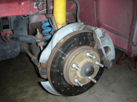1998 Ford Crown Victoria - Close-up of a front brake assembly showing a drilled rotor, five wheel studs, caliper, blue coil spring and yellow strut on a red-bodied ca…