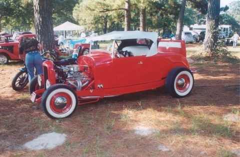 2002 Ford Crown Victoria - A red hot rod roadster is parked outdoors with other vehicles and people in the background.