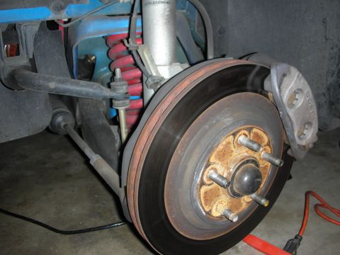 1998 Ford Crown Victoria - Close-up of a brake rotor and rusty wheel hub with five studs, alongside red coil springs and suspension control arms under a red car.