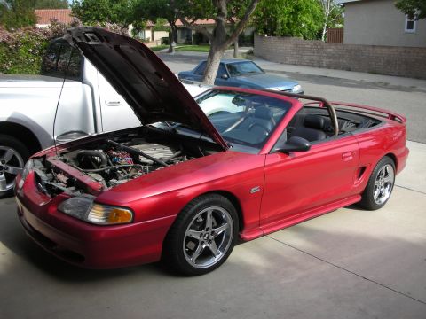 1998 Ford Crown Victoria - Red Ford Mustang GT convertible with the hood raised and top down, parked in a residential driveway beside other cars.