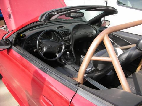 1998 Ford Crown Victoria - Interior of a red Mustang convertible showing the dashboard, manual shifter, leather seats and a tan roll bar with top down.