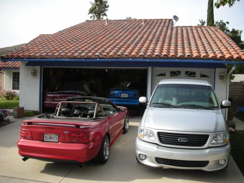 1998 Ford Crown Victoria - Red Mustang convertible and a silver Ford pickup parked outside a Spanish tile-roof home, with a blue truck visible in the garage.
