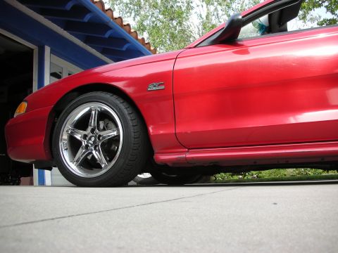 1998 Ford Crown Victoria - Low ground-level front-quarter view of a red Mustang on a chrome five-spoke wheel, parked beside a blue-trimmed garage.