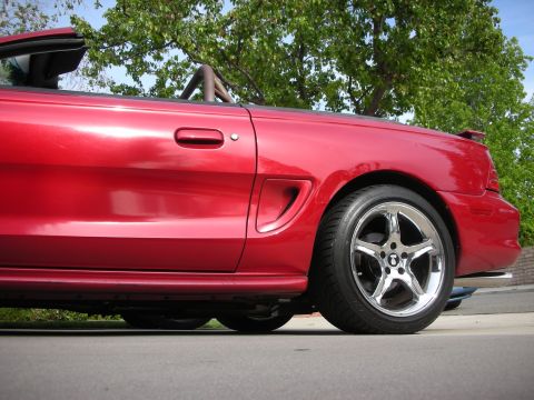 1998 Ford Crown Victoria - Rear-quarter view of a red Mustang convertible with top down on a chrome five-spoke wheel, parked under a tree.