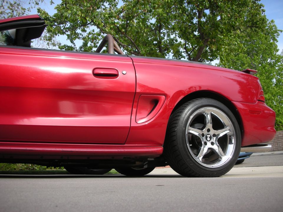 Rear quarter of red Mustang convertible on chrome wheel under a tree