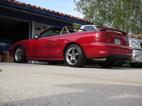 1998 Ford Crown Victoria - Rear side view of a red Mustang GT convertible with top down and tan roll bar, parked in a driveway by a garage.