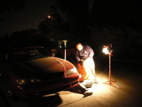 1998 Ford Crown Victoria - Nighttime driveway scene with two people working near the front of a red convertible, illuminated by a portable work light.