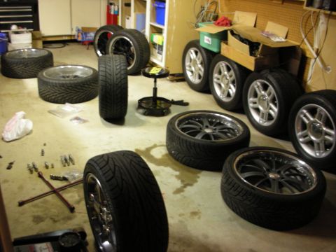 1998 Ford Crown Victoria - Garage floor covered with multiple alloy and chrome wheels mounted on low-profile tires, plus a lug wrench and scattered tools.