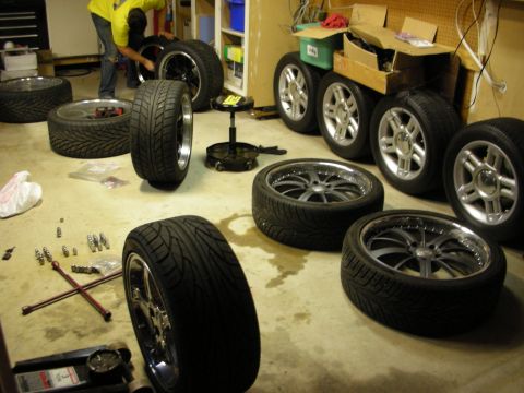 1998 Ford Crown Victoria - person in a yellow shirt works among several alloy and chrome wheels with low-profile tires spread across a garage floor.