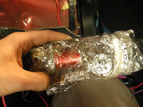 1998 Ford Crown Victoria - hand holds a plastic-wrapped Kicker audio crossover with red coil winding, resting on a knee amid car wiring.